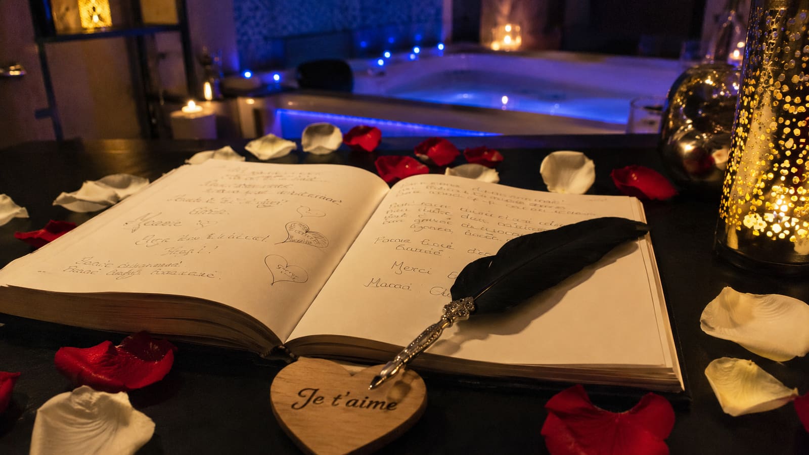 Livre d'or ouvert avec une plume, entouré de pétales de rose et d'un cœur en bois gravé « Je t'aime », jacuzzi éclairé en bleu en arrière-plan