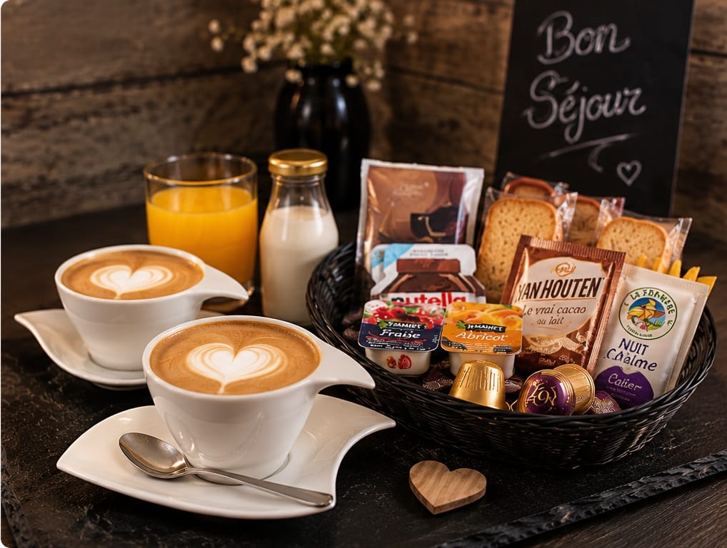 Deux cafés au lait au décor de cœur sur des soucoupes blanches, un verre de jus d&rsquo;orange et une bouteille de lait, et une corbeille garnie de biscottes, de dosettes de cacao, de confiture, de miel et de pâte à tartiner, devant une ardoise où est écrit « Bon séjour ».