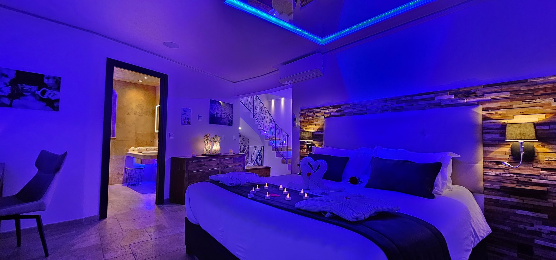 La chambre en vue large sous éclairage bleu, le lit avec des serviettes pliées en cœur et des bougies, l’escalier et l’ouverture sur la salle de bain
