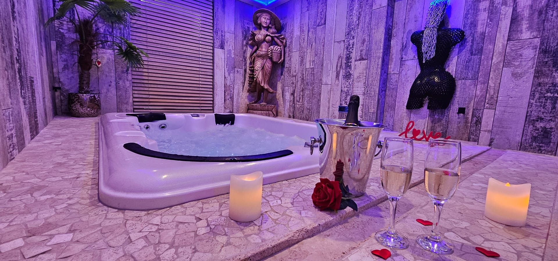 Le jacuzzi sous éclairage violet, un seau à champagne, des bougies et des pétales de rose rouges posés au bord