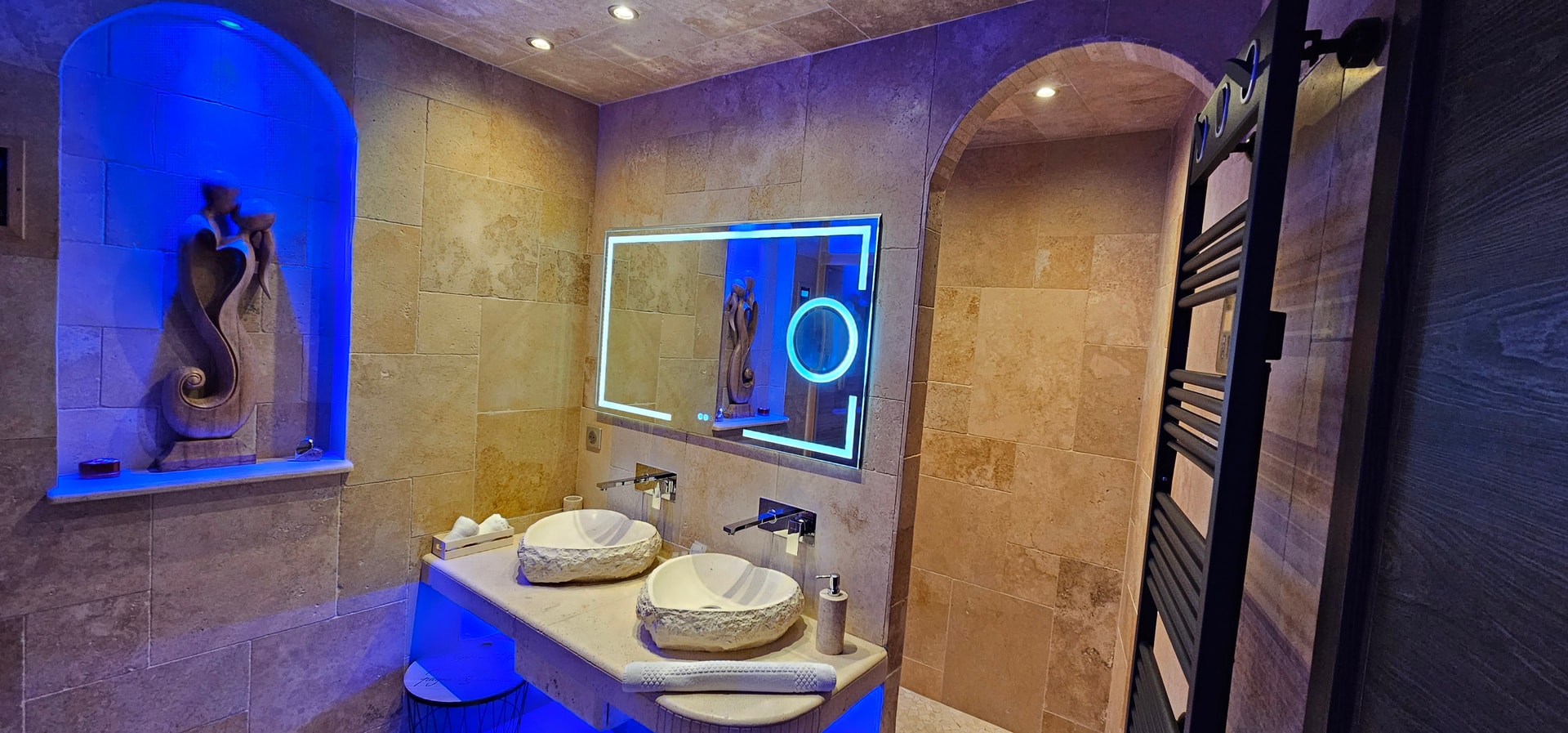 La salle de bain en vue large : deux vasques de pierre, miroir lumineux, miroir grossissant et sèche-serviettes